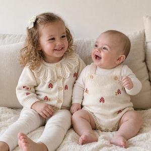 Pode incluir: Um cardigã de malha cor de creme com um grande colarinho de folhos e bordados de cogumelos vermelhos, combinado com leggings creme. Ao lado, um macacão de malha creme com bordados de cogumelos vermelhos e botões de madeira sobre uma camisa.