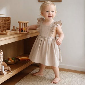 Könnte beinhalten: Ein beiges ärmelloses Kleid mit quadratischem Ausschnitt und gerüschten Schulterträgern. Das Kleid verfügt über einen gerafften Rock und ein offenes Seitendesign mit Bindebändern am Rücken, gefertigt aus einem einfarbigen Stoff mit sichtbarer Textur.
