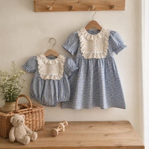 May include: A blue and white gingham dress and a matching romper, both featuring short puff sleeves and a cream-colored lace-trimmed bib collar. The garments are displayed on wooden hangers against a neutral wall, showcasing a coordinated set.