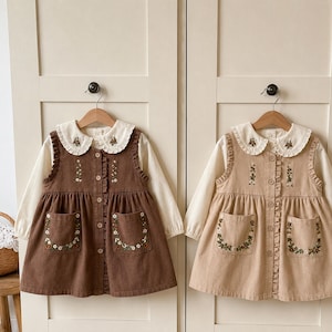 May include: Two sleeveless dresses with button-down fronts, ruffled armholes, and two front pockets. One dress is dark brown and the other is light tan. Both feature floral embroidery on the pockets and collars, paired with long-sleeved white shirts.