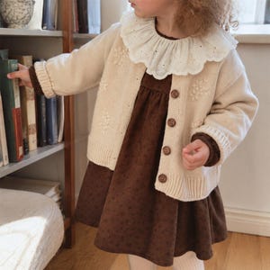 Puede incluir: Una chaqueta de punto color crema con bordados florales, dobladillo acanalado y cuatro botones marrones, sobre un vestido marrón oscuro con un patrón sutil. La chaqueta presenta un cuello grande de encaje blanco con ojales festoneados.