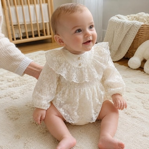 May include: An off-white long-sleeve romper featuring an intricate floral lace overlay, a ruffled collar, and two small pearl-like buttons at the neckline. The garment has long sleeves with gathered cuffs and a full, layered lace skirt design.