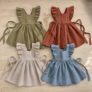 Puede incluir: Cuatro vestidos sin mangas en verde oliva, naranja terracota, beige y azul polvoriento. Cada vestido presenta un escote cuadrado, tirantes anchos con volantes, una falda fruncida y lazos laterales largos que se atan alrededor de la cintura.