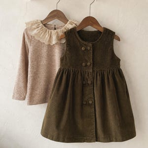 Peut inclure: Une robe sans manches en velours côtelé vert olive avec une encolure ronde et six boutons recouverts de tissu assorti sur le devant, associée à un haut beige chiné à manches longues avec un large col à volants en dentelle crème.