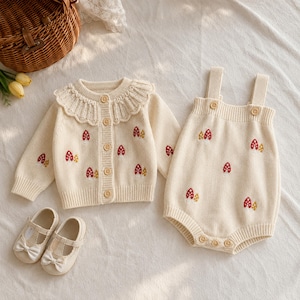 Pode incluir: Um cardigã de malha cor de creme com gola de folhos e fecho de botões, combinado com um macacão sem mangas a condizer com um padrão de cogumelos em vermelho e amarelo. Inclui um par de sapatos Mary Jane cor de creme com laços.