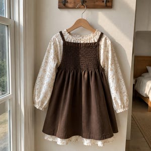 Könnte beinhalten: Ein dunkelbraunes Trägerkleid mit gesmoktem Oberteil über einer cremefarbenen Langarmbluse mit zartem braunem Blumenmuster. Die Bluse hat einen Rüschenkragen und elastische Bündchen, während das Trägerkleid einen weit ausgestellten Rock besitzt.