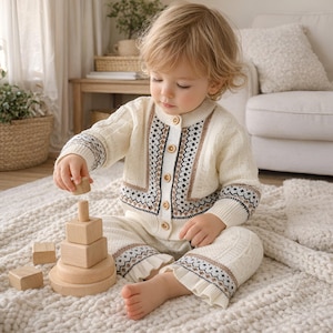 Peut inclure: Un bébé portant un cardigan en tricot crème et un pantalon assorti avec un motif géométrique. La tenue a des boutons et des bordures en bois. Le bébé joue avec un jouet d'empilage en bois sur un tapis blanc.