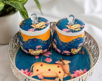 May include: Two decorative jars with lids and a serving tray. The jars have a blue and orange design with a cow and floral pattern. The tray is silver with a blue cow and lotus flower design. A plant is in the background.