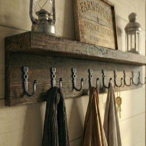 May include: A rustic wooden shelf with a row of metal hooks below. The shelf holds a vintage lantern and a sign that reads "Farmhouse Vintage Sticks". Scarves and keys hang from the hooks, creating a functional and decorative storage solution.