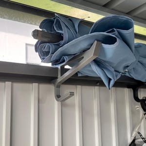 Könnte beinhalten: Ein hellblauer, zusammengeklappter Regenschirm liegt auf einem silbernen Metallhaken, der an einer vertikal gerippten Wandplatte befestigt ist. Der Haken hat eine flache Oberseite und ein gebogenes Unterteil. Rechts ist ein schwarzer Haken sichtbar.