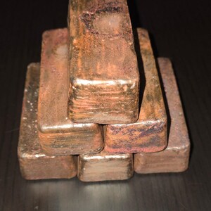 Op de afbeelding: Een stapel van zes rechthoekige, koperkleurige metalen staven. De staven hebben een ruw, getextureerd oppervlak met donkerdere oxidatiegebieden. Ze zijn in een piramidevorm gerangschikt, met vier staven als basis.