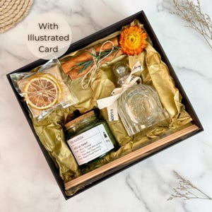 May include: A black gift box containing a clear glass diffuser bottle, a small green jar, dried orange slices, cinnamon sticks, and a dried orange flower. The box is lined with gold tissue paper.
