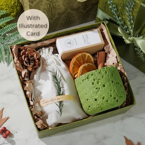 May include: A green gift box containing a selection of items. It includes a white drawstring bag, a green textured container, dried orange slices, a pine cone, and a small box. The box is labelled "With Illustrated Card."
