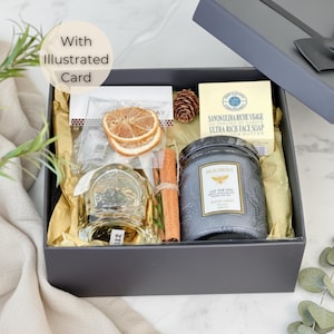 Può includere: Una scatola regalo grigia con coperchio parzialmente aperto, riempita con una candela, sapone, una bottiglia, fette d'arancia essiccate e un bastoncino di cannella. La candela ha il testo "JUST FOR YOU". La scatola contiene anche una carta illustrata.