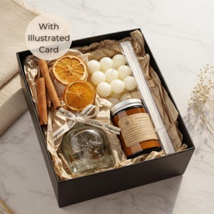 Op de afbeelding: Zwarte geschenkdoos gevuld met een schedelvormige glazen fles, een potkaars, witte bolkaarsen, kaneelstokjes, gedroogde sinaasappelschijfjes en rietverspreiders. De doos bevat een lint met tekst en een kaart met "With Illustrated Card."