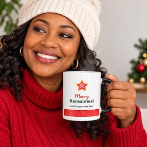 May include: A white ceramic mug with a black handle featuring a red star graphic with a white snowflake pattern, the text "Merry Keresimesi and Happy New Year" in red and green, and a red horizontal band with small gold dots at the bottom.