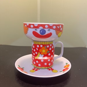 May include: A clown-themed ceramic teacup, bowl, and saucer set. The teacup and bowl feature a clown face design with a red nose and a polka-dot pattern. The saucer has floral accents. A fun, vintage-inspired kitchenware set.
