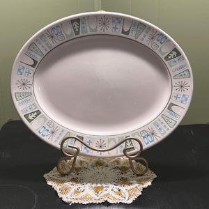 May include: An oval, white ceramic platter with a decorative border featuring geometric shapes in blue, green, and black. The platter is displayed on a gold-toned metal stand over a white and gold doily. A vintage serving dish.