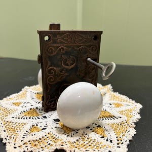 May include: An antique, ornate metal lock with a key inserted, resting on a white and gold doily. Two white, egg-shaped objects are in the foreground. The lock has intricate scrollwork and a dark, aged finish.