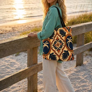 May include: A tote bag featuring a repeating pattern of dark blue palm trees and intricate orange and cream floral motifs. The bag has two black shoulder straps and a rectangular shape, displaying a detailed tile-inspired design on the fabric.