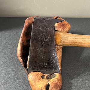 May include: A metal axe head with a dark, weathered, and pitted surface texture attached to a light-colored wooden handle. The axe is resting against a piece of natural, light brown, irregularly shaped wood with a hollow, organic, and knotty texture.