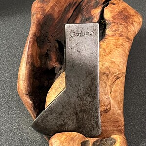 May include: A dark gray metal axe head with the word Norlund stamped on the upper portion. The axe head is positioned against a piece of light brown, textured, organic-shaped wood with dark brown grain patterns and natural knots throughout.