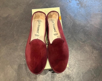 May include: A pair of burgundy velvet slip-on shoes with a rounded toe. The interior insoles are cream-coloured and feature the text Gio Dreams, Prodotto Italiano, and the size 38. The shoes are resting on a light-coloured cardboard box.