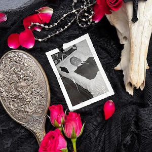 May include: A still life arrangement featuring a vintage hand mirror, red roses, and a black and white photograph. A skull with a cross and a beaded necklace is also present. The items are arranged on a black lace fabric, with scattered rose petals.