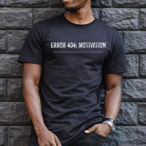 May include: Black t-shirt with the text "ERROR 404: MOTIVATION" in white, with the text "resource temporarily unavailable" below it. The shirt is worn by a person leaning against a dark gray brick wall.