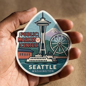 Puede incluir: Una pegatina troquelada con una ilustración del paisaje urbano de Seattle. Muestra la Space Needle, una noria, un ferry y el texto PUBLIC MARKET CENTER, SEATTLE, WASHINGTON en rojo y blanco sobre un fondo azul y verde.