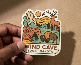 May include: A die-cut sticker featuring a colorful illustration of a landscape with a cave entrance, a cactus, a deer, and a bison. The scene includes mountains and a sun under a clear sky. The text reads WIND CAVE SOUTH DAKOTA in bold letters.