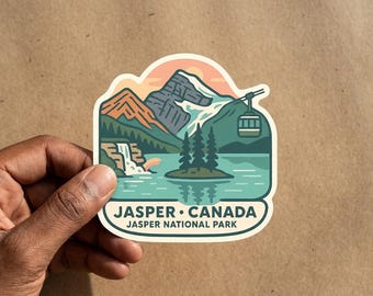 May include: A die-cut sticker featuring a graphic illustration of Jasper National Park in Canada. The design displays mountains, a waterfall, a lake with an island of pine trees, and a cable car. The text reads JASPER CANADA and JASPER NATIONAL PARK.