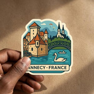 Puede incluir: Una pegatina troquelada con una ilustración estilizada de Annecy, Francia, con un castillo, un puente, un cisne en el agua y parapentes en el cielo. El texto ANNECY • FRANCE está impreso en negro en la parte inferior del borde crema.