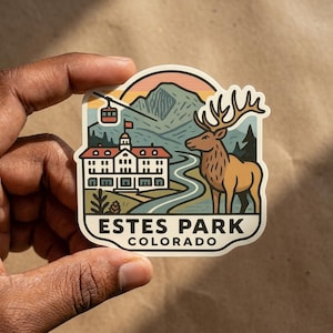 May include: A die-cut sticker featuring a stylized illustration of Estes Park, Colorado. The design includes a large elk, a historic hotel building, a mountain range, a cable car, and a winding river. The text ESTES PARK COLORADO is printed at the bottom.