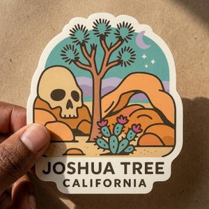 Op de afbeelding: Een gestanste sticker met een woestijnlandschap met een Joshua tree, een menselijke schedel, oranje rotsformaties en een cactus met roze bloemen. De lucht is blauwgroen met een maansikkel en sterren. De tekst luidt JOSHUA TREE CALIFORNIA.