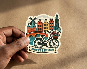 Puede incluir: Una pegatina troquelada con una colorida ilustración de Ámsterdam que muestra un molino de viento, casas de canal, un puente y una bicicleta con una cesta de tulipanes rojos. La palabra AMSTERDAM aparece en letras azules sobre una cinta inferior.