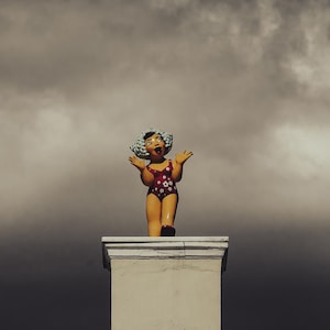 Puede incluir: Figura de cerámica de una mujer con un bañador rojo con estampados florales blancos y un peinado rizado azul. La figura está de pie sobre un pilar blanco contra un cielo nublado.