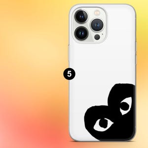 Op de afbeelding: Witte telefoonhoes met een zwart hartvormig ontwerp met twee ogen. De hoes heeft een transparante rand en een cameramodule met drie lenzen. De achtergrond is een verloop van geel en oranje.