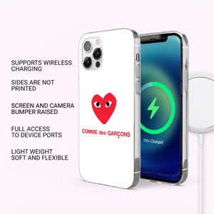 May include: A white phone case featuring a red heart design with eyes and the text "COMME des GAR&Ccedil;ONS". The phone case is shown with a phone displaying a charging screen. The case supports wireless charging and has full access to device ports.