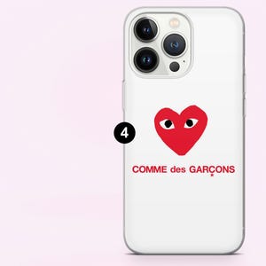 May include: White iPhone case featuring a red heart with eyes and the text "COMME des GAR&Ccedil;ONS" in red. The case has a clear border and covers the phone's back and sides.