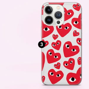 May include: A clear phone case featuring a pattern of red hearts with black eyes on a white background. The case has a clear border and covers the back and sides of the phone. The phone's camera lenses are visible.