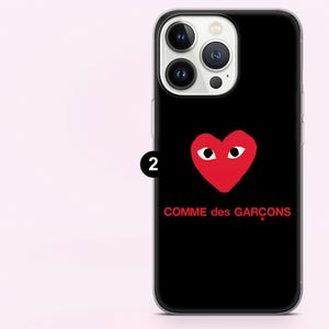 May include: Black phone case featuring a red heart with eyes and the text "COMME des GAR&Ccedil;ONS" in red. The case has a clear border and is designed for a smartphone.