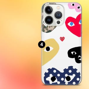 Op de afbeelding: Transparante telefoonhoes met een witte achtergrond met kleurrijke hart- en oogontwerpen. De hoes heeft een rood hart met blauwe ogen, een geel hart met een oog en een zwart hart met ogen. Er is ook een klein rood hart aanwezig.