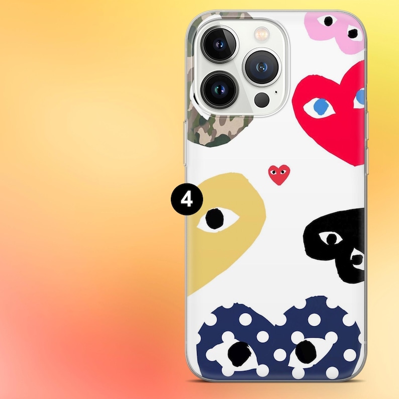 Op de afbeelding: Transparante telefoonhoes met een witte achtergrond met kleurrijke hart- en oogontwerpen. De hoes heeft een rood hart met blauwe ogen, een geel hart met een oog en een zwart hart met ogen. Er is ook een klein rood hart aanwezig.