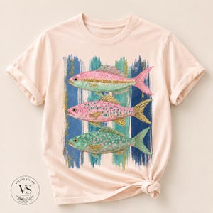 Pode incluir: Uma t-shirt de cor p&ecirc;ssego claro com uma estampa gr&aacute;fica de tr&ecirc;s peixes empilhados verticalmente. Os peixes s&atilde;o representados em tons de rosa, azul-petr&oacute;leo e detalhes em glitter dourado, sobre um fundo de pinceladas verticais azuis.