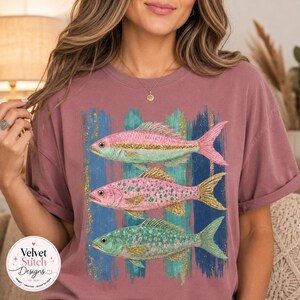 Pode incluir: Uma t-shirt de manga curta em rosa seco com um gr&aacute;fico de tr&ecirc;s peixes empilhados verticalmente. Os peixes s&atilde;o representados em tons de rosa e azul-petr&oacute;leo com detalhes em glitter dourado sobre um fundo de pinceladas azuis.
