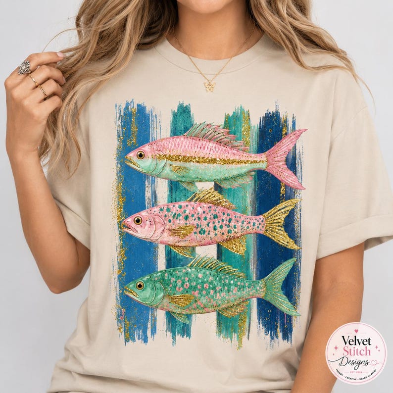 Pode incluir: Uma t-shirt bege com um gr&aacute;fico de tr&ecirc;s peixes empilhados em tons de rosa, verde e dourado. Os peixes est&atilde;o sobrepostos a pinceladas verticais de tinta azul e verde com detalhes de glitter dourado. O design est&aacute; posicionado no peito.