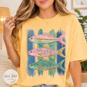 Pode incluir: Uma t-shirt amarela com uma estampa gr&aacute;fica de tr&ecirc;s peixes empilhados verticalmente. Os peixes s&atilde;o representados em tons de rosa, verde e com detalhes de glitter dourado, sobre um fundo de pinceladas verticais azuis e verde-azuladas.