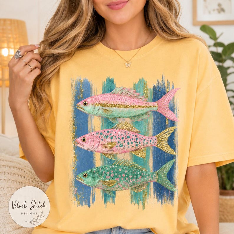 Pode incluir: Uma t-shirt amarela com uma estampa gr&aacute;fica de tr&ecirc;s peixes empilhados verticalmente. Os peixes s&atilde;o representados em tons de rosa, verde e com detalhes de glitter dourado, sobre um fundo de pinceladas verticais azuis e verde-azuladas.