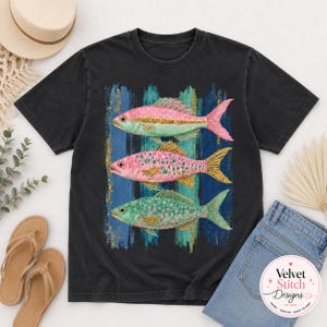 Pode incluir: Uma t-shirt preta de manga curta com uma estampa gr&aacute;fica de tr&ecirc;s peixes empilhados verticalmente. O peixe de cima &eacute; rosa, o do meio &eacute; rosa com pontos dourados e o de baixo &eacute; verde-azulado com pontos rosa, sobre um fundo de pinceladas azuis.